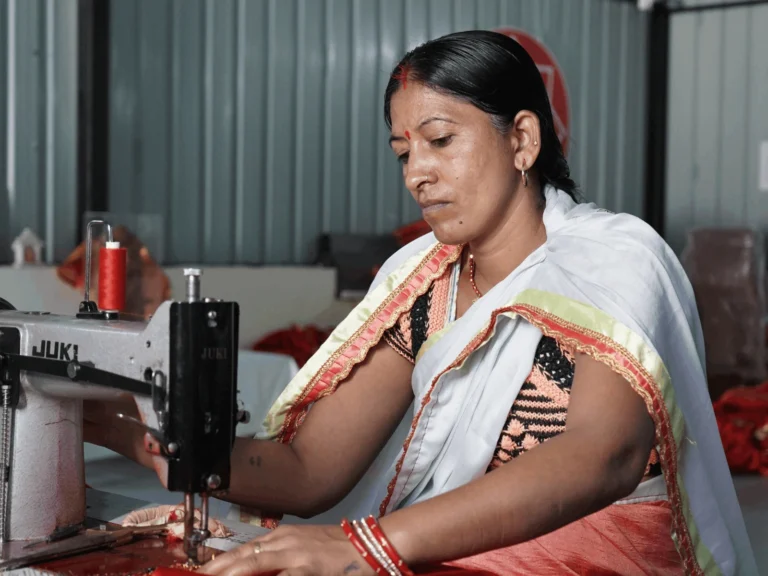 A woman dressed in a traditional sari sitting at a JUKI sewing machine, focused on her stitching work in a workshop.