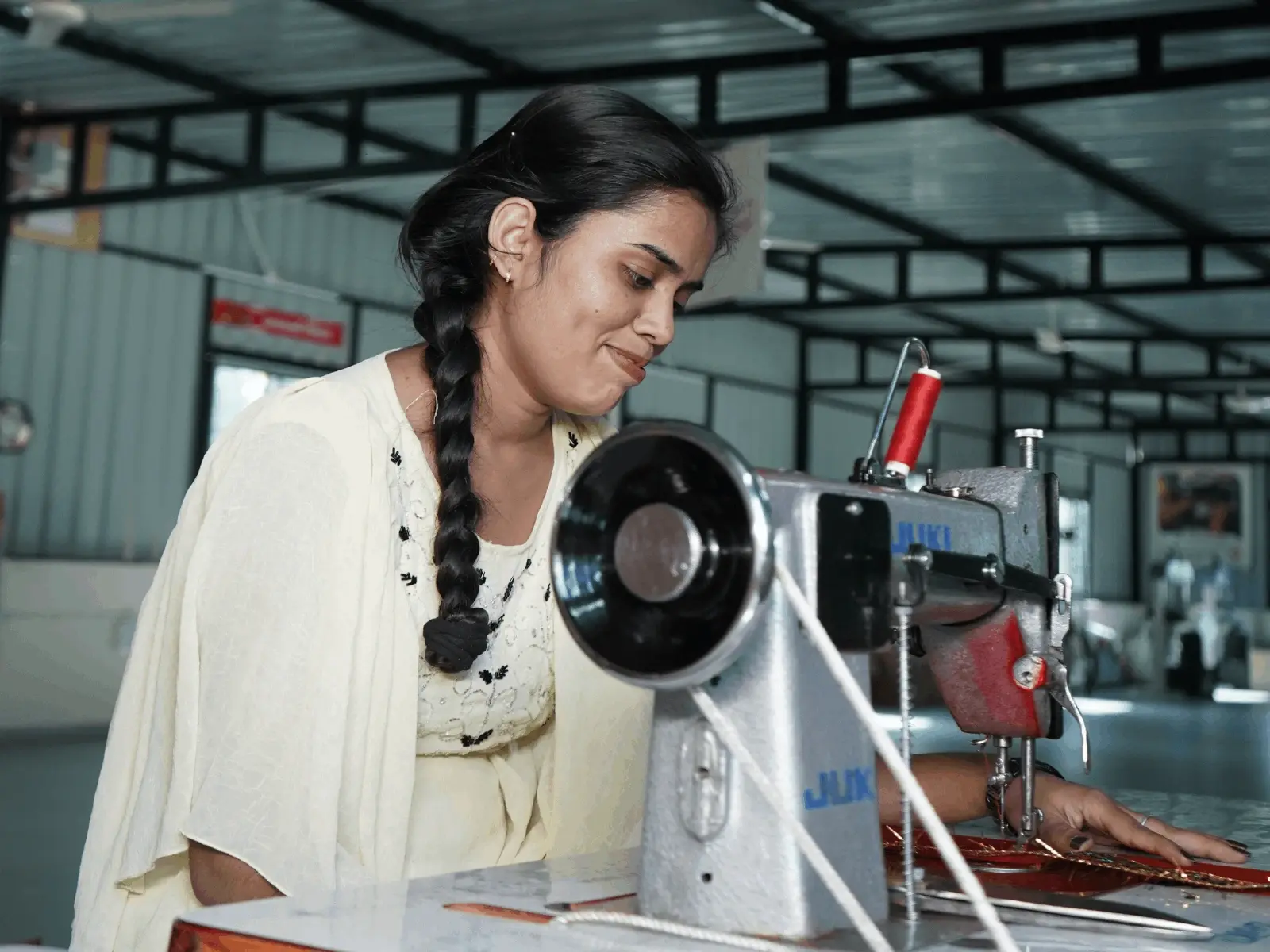 A smiling young woman with a long side braid working at a professional JUKI sewing machine in a workshop.
