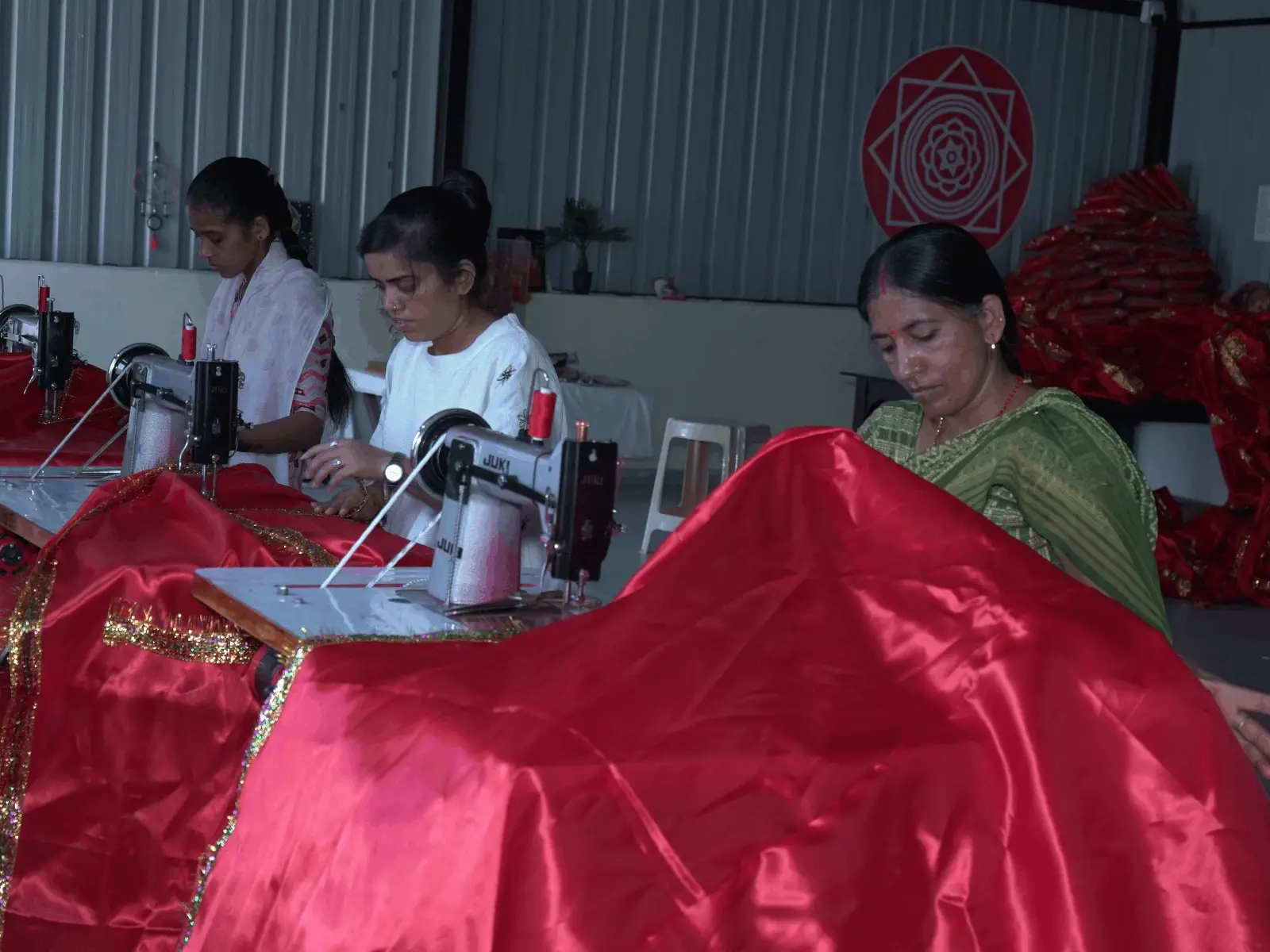 Three women working at sewing machines, stitching red fabric with gold trim in a textile workshop.