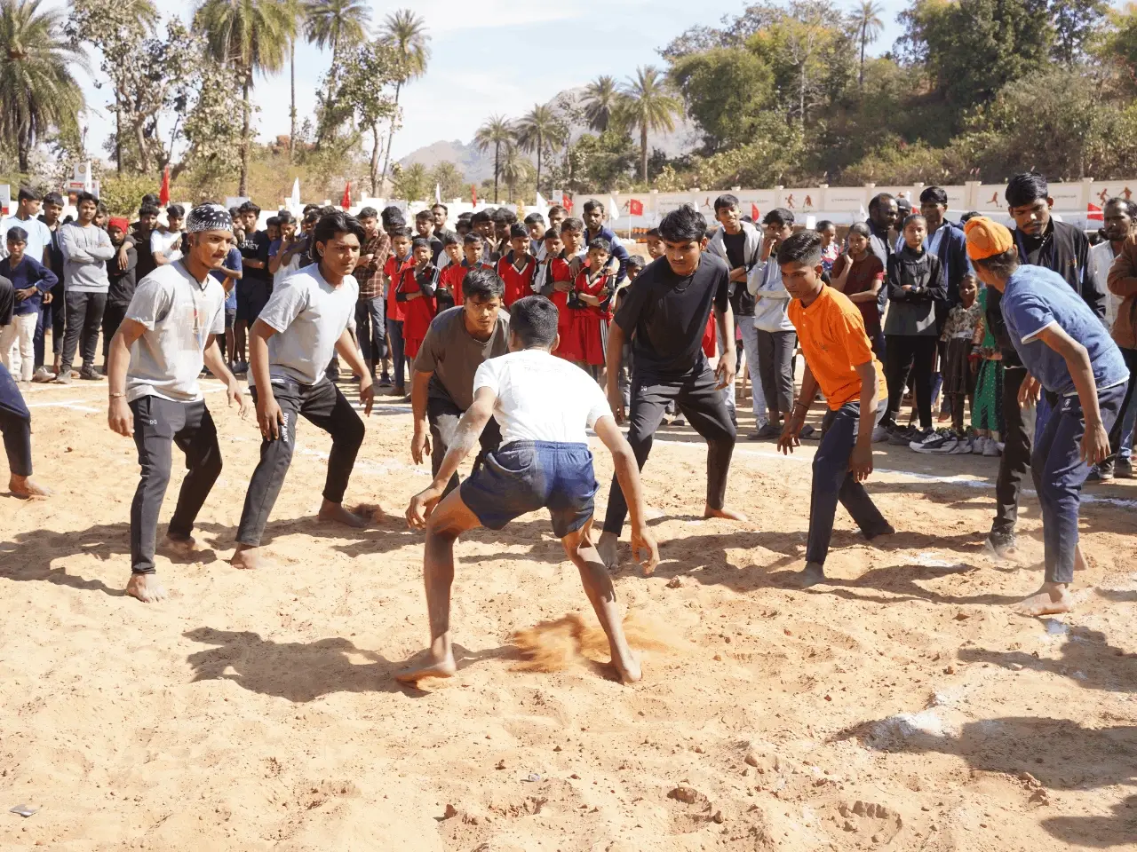 A group of young men playing a competitive game of kabaddi on a sandy court, with a crowd of spectators in the background.